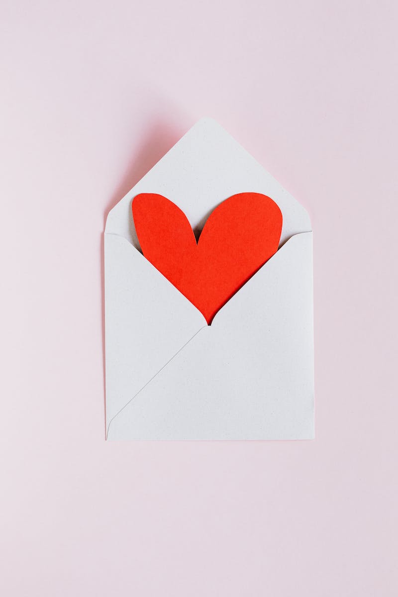 Top view of opened white envelope with handmade red paper heart placed against pink background as gift for Valentine day