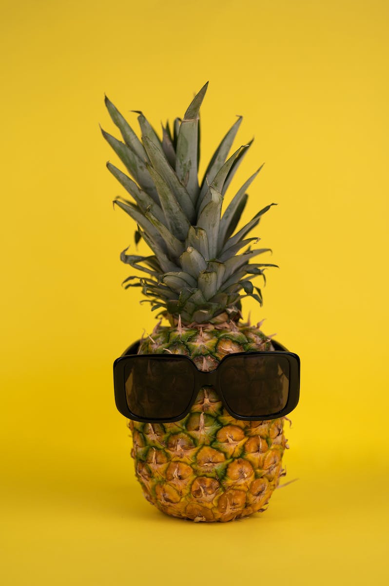 Whole ripe pineapple with green crown and brown skin and modern black sunglasses placed on yellow background in light studio