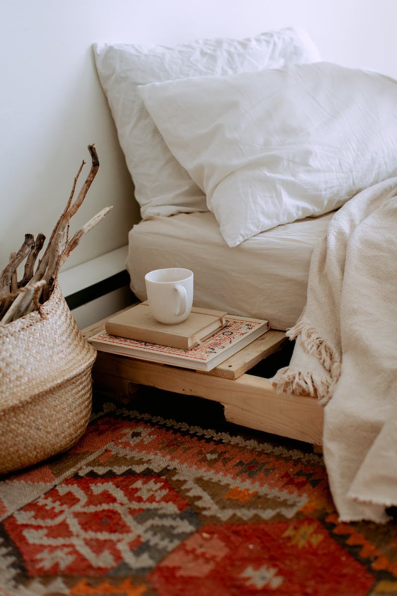 Comfortable cozy bedroom with bed wooden shelves with book and cup while wicker basket with sticks placed on ethnic styled rug