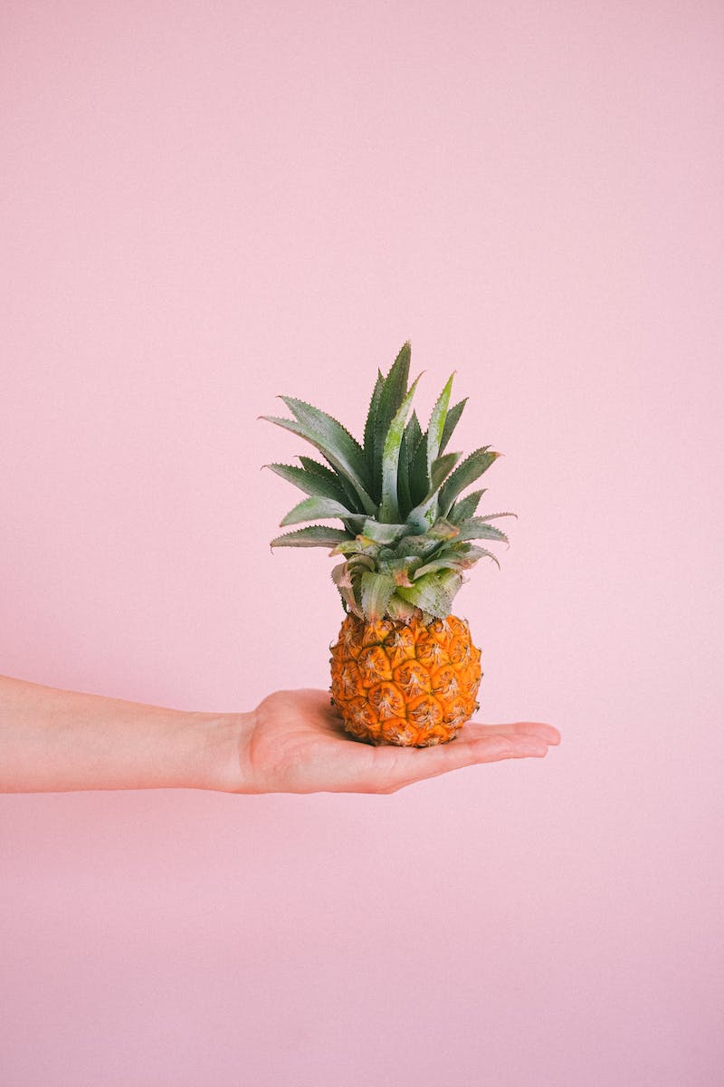 Crop anonymous person demonstrating small ripe pineapple on hand against light pink background in modern studio