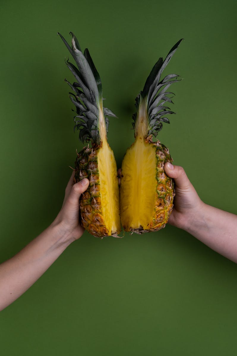 Crop faceless people with halves of ripe pineapple with green leaves in hands standing on green background in light studio