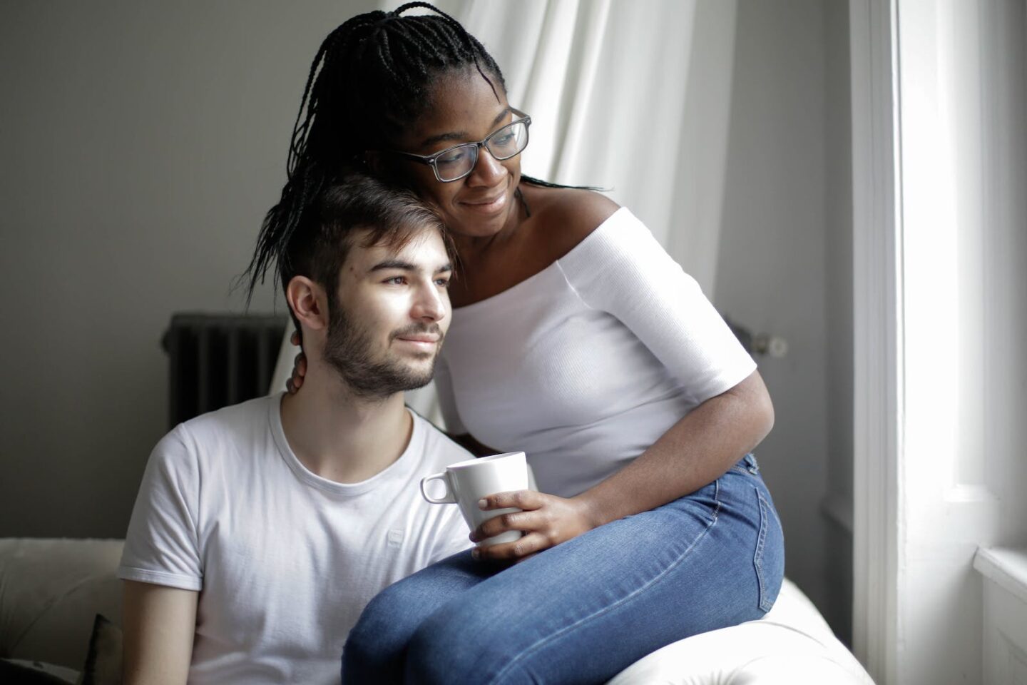 Positive young multiracial couple in casual clothes sitting on comfortable sofa with cup and looking away through window in modern living room