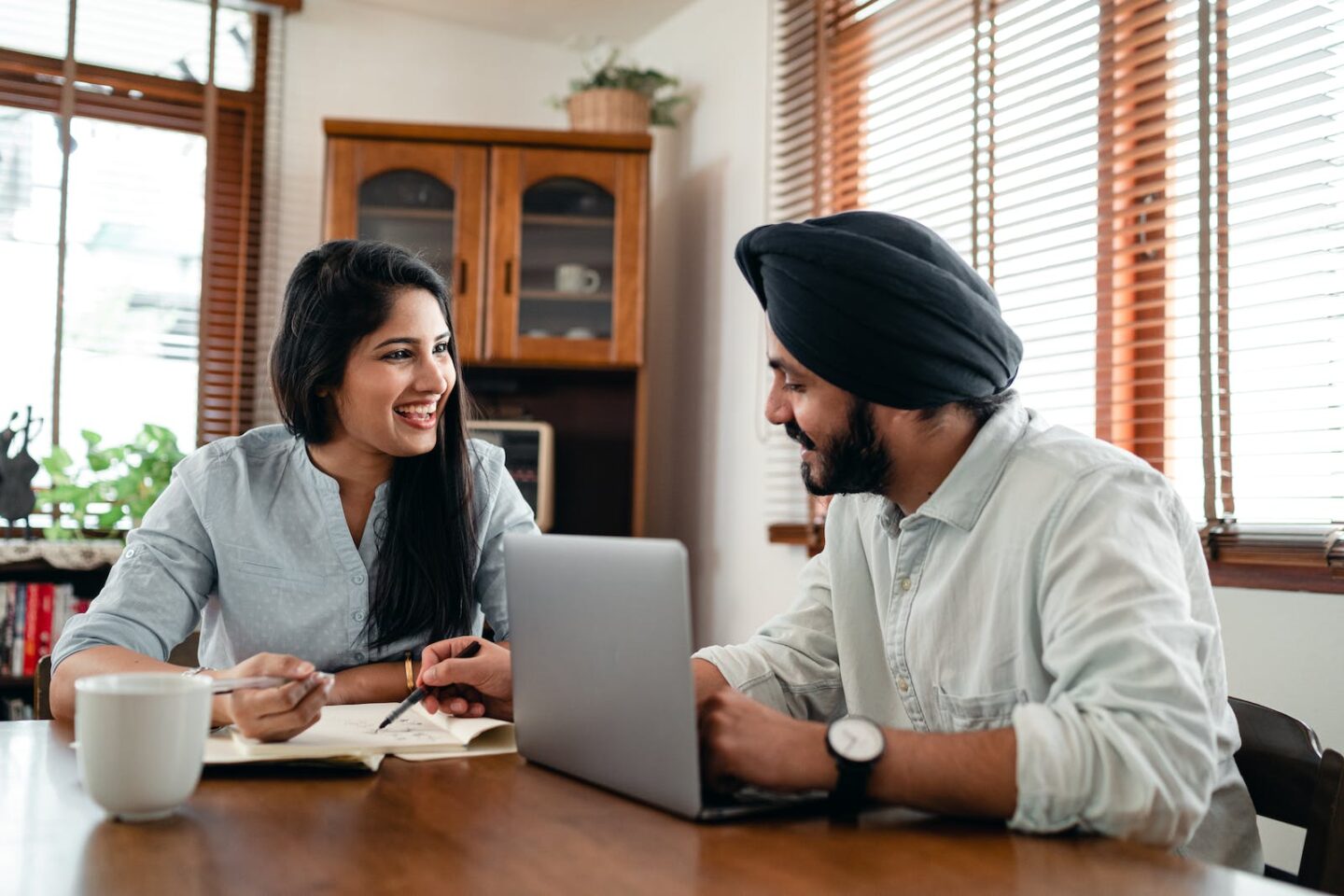 Laughing Indian woman with husband sitting at table and discussing project while working together with laptop and notebook at home